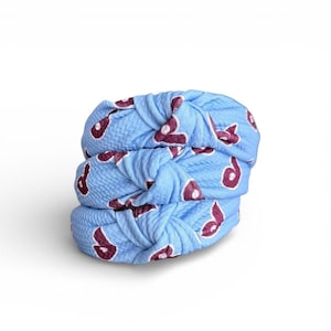 May include: Three light blue knotted headbands stacked on top of each other. Each headband has a repeating pattern of a maroon-colored design. The headbands are made of a textured fabric.