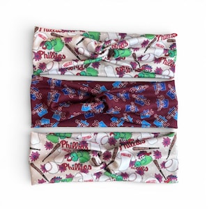 May include: Three Philadelphia Phillies headbands. Two are cream-colored with baseballs, bats, flowers, and the Phillies logo. The other is maroon with the Phillies logo in blue and white.