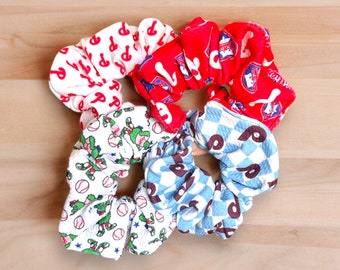 May include: Four scrunchies made of fabric with different patterns. One scrunchie is red with white and blue baseball-themed designs. Another scrunchie is white with green baseball players. A third scrunchie is white with red and blue "P"s. The last scrunchie is blue and white with brown "P"s.