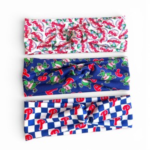 May include: Three headbands with a knotted design. One is white with red and blue Phillies logos, another is blue with green and red cartoon characters, and the third is blue and white checkered with red "P" logos.