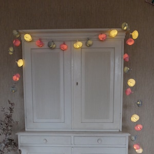 Może przedstawiać: Biała drewniana szafka z dwojgiem drzwi i dwoma szufladami. Szafka ma ozdobną listwę wokół górnej i dolnej części. Nad szafką wisi sznur światełek z papierowymi lampionami w odcieniach różowego, żółtego i zielonego.