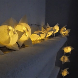 May include: A string of white paper lanterns with warm yellow lights inside. The lanterns are hanging from a white wall and are arranged in a row.