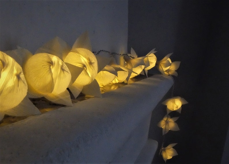 May include: A string of white paper lanterns with warm yellow lights inside. The lanterns are hanging from a white wall and are arranged in a row.