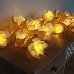 Peut inclure: Une guirlande de lanternes en papier en forme de fleurs, avec des lumières blanches chaudes à l'intérieur, suspendue à une table en bois.