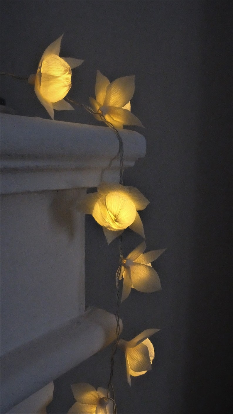 May include: A string of white paper flower lights with warm white LED bulbs. The lights are strung along a white cord and are hanging from a white wall.