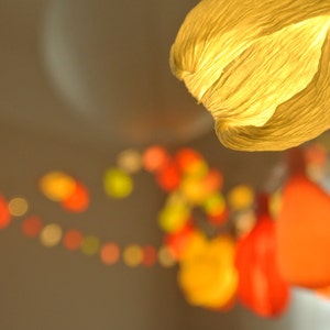 Puede incluir: Una cadena de farolillos de papel de varios colores, incluyendo naranja, amarillo y rojo, cuelgan del techo. Los farolillos están hechos de papel fino y arrugado y están iluminados desde el interior, creando un ambiente cálido y acogedor.