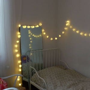 Puede incluir: Una cama con estructura de metal blanco, colchón blanco y una manta blanca. La cama está decorada con una cadena de luces blancas.