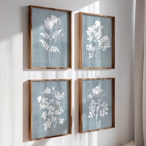 May include: Four rectangular wall art pieces in wooden frames, each featuring a white botanical illustration of flowers and leaves set against a solid, muted blue background. The prints are arranged in a two-by-two grid on a plain white wall.