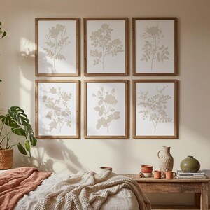 May include: A set of six framed botanical wall art prints featuring beige floral illustrations on white backgrounds, arranged in two rows of three. The prints are displayed above a bed with terracotta bedding and a chunky knit throw blanket.