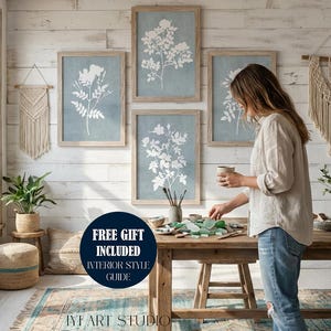 May include: A set of four framed botanical prints featuring white floral silhouettes on a muted blue background, displayed on a white wood-paneled wall. The artwork is accompanied by two cream-colored macrame wall hangings and a wooden table.