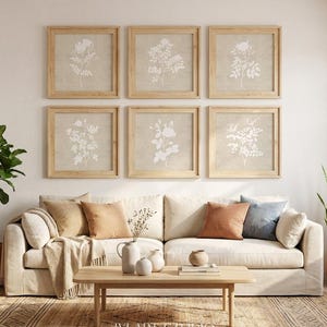 May include: Six square framed wall art pieces featuring white botanical illustrations on a beige background, arranged in two rows above a beige sofa. The text IYI ART STUDIO is visible at the bottom centre of the image, below the wooden coffee table.