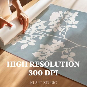 May include: A large square print featuring a white botanical rose branch silhouette against a muted blue background. The print includes the text HIGH RESOLUTION 300 DPI and IYI ART STUDIO. The paper is partially rolled up on a wooden surface.