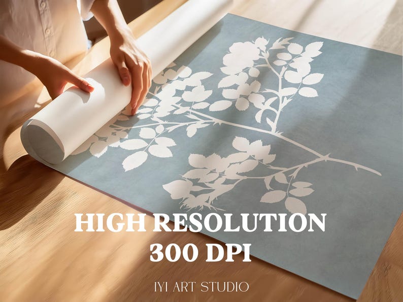 May include: A large square print featuring a white botanical rose branch silhouette against a muted blue background. The print includes the text HIGH RESOLUTION 300 DPI and IYI ART STUDIO. The paper is partially rolled up on a wooden surface.