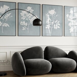 May include: A dark gray velvet sofa with a rounded, organic silhouette featuring two main seating sections and curved side bolsters. Behind the sofa are four framed wall art prints displaying white botanical floral silhouettes on a dusty blue background.