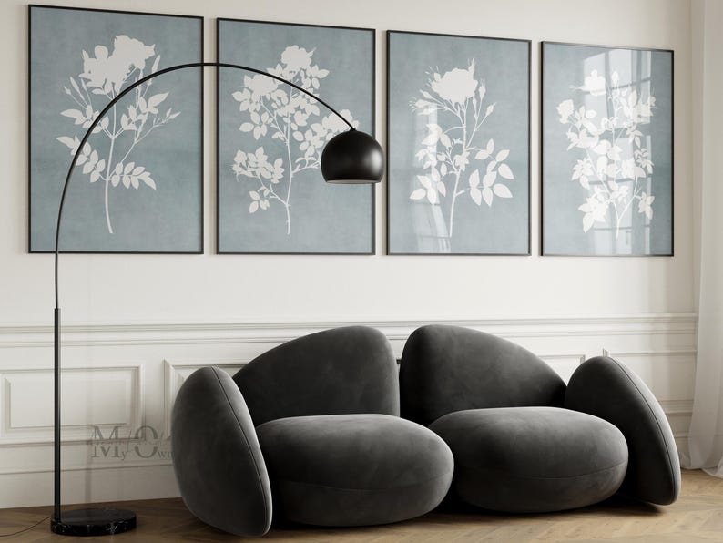 May include: A dark gray velvet sofa with a rounded, organic silhouette featuring two main seating sections and curved side bolsters. Behind the sofa are four framed wall art prints displaying white botanical floral silhouettes on a dusty blue background.