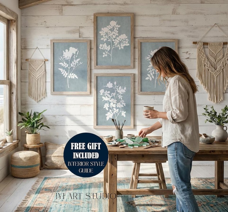 May include: A set of four framed botanical prints featuring white floral silhouettes on a muted blue background, displayed on a white wood-paneled wall. The artwork is accompanied by two cream-colored macrame wall hangings and a wooden table.