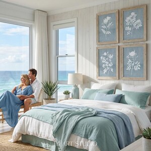 May include: A bedroom featuring a bed with white and light blue bedding, two light blue table lamps, and four framed botanical prints on a white paneled wall. A wooden bench sits near a large window overlooking the ocean.