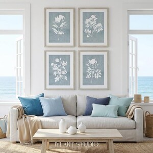 May include: A set of four framed wall art prints featuring white botanical floral silhouettes against a muted blue background, arranged in a grid pattern above a light-colored sofa with blue and beige throw pillows and a wooden coffee table.