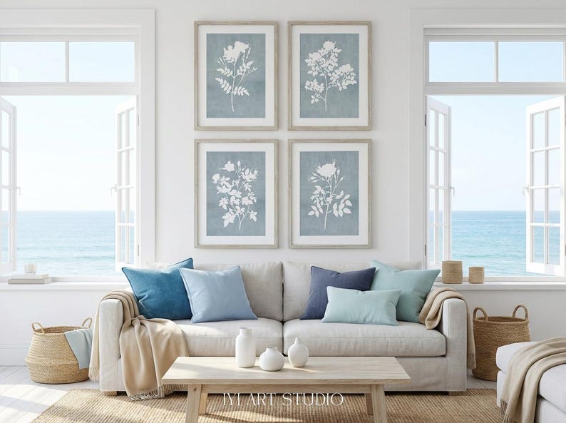 May include: A set of four framed wall art prints featuring white botanical floral silhouettes against a muted blue background, arranged in a grid pattern above a light-colored sofa with blue and beige throw pillows and a wooden coffee table.