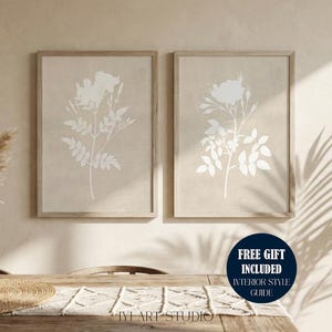 May include: Two framed vertical wall art prints featuring white botanical floral silhouettes on a light beige background. The prints are displayed in light wood frames. A dark circular badge in the corner contains the text FREE GIFT INCLUDED INTERIOR STYLE GUIDE.
