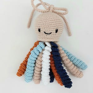 Op de afbeelding: Een handgemaakt gehaakt octopus speelgoed met een beige hoofd en een lachend gezicht. De octopus heeft acht kleurrijke tentakels in tinten oranje, blauw, wit en marine. Een beige strik is bevestigd aan de bovenkant van het hoofd.
