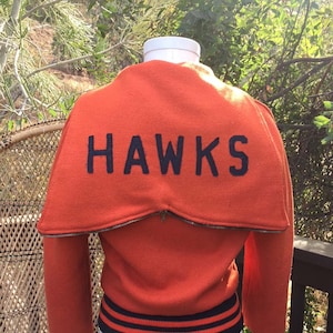 Pode incluir: Casaco de beisebol laranja com acabamento em listras pretas e brancas e a palavra "HAWKS" bordada nas costas.