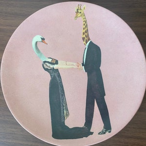 Puede incluir: Un plato de cerámica rosa con un diseño caprichoso que presenta una jirafa con esmoquin tomando la mano de un cisne con un vestido negro.