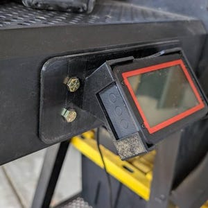 May include: A black digital display unit with a red border mounted to a black metal frame using two silver-colored bolts. The device features a rectangular screen and three circular buttons on the side, attached to a perforated metal surface.