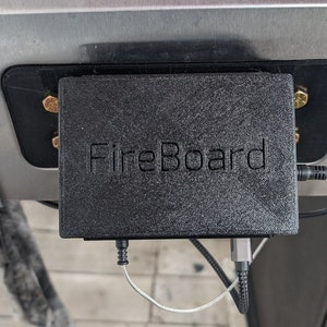 May include: A black FireBoard device is mounted on a stainless steel grill. The device is rectangular with the word "FireBoard" embossed on the front. Cables and connectors extend from the device.