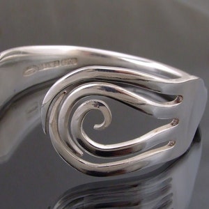 May include: A silver bracelet made from a repurposed fork. The design incorporates a decorative swirl on the front, with the fork tines integrated into the bracelet's structure. The bracelet has a polished, reflective finish.