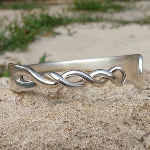 May include: A silver bracelet with a braided design. The bracelet is made from a single piece of metal that is twisted and shaped into a continuous braid.