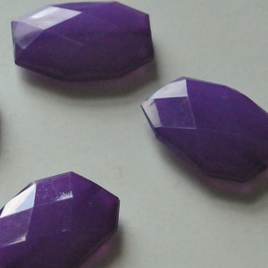 May include: Close-up of several faceted, elongated purple beads. The beads have a geometric shape with multiple flat surfaces that reflect light. The color is a deep, rich purple, and the beads appear to be made of a translucent material.