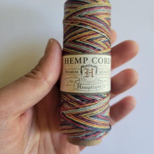 Puede incluir: Un carrete de cuerda de cáñamo multicolor, con una etiqueta que dice "Hemp Cord" y "Hemptique". La cuerda es una mezcla de colores que incluyen rojo, amarillo, azul y marrón. La etiqueta también dice "Premium Quality".