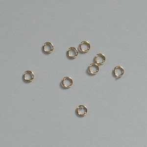 May include: A scattering of ten small, gold jump rings on a light gray surface.