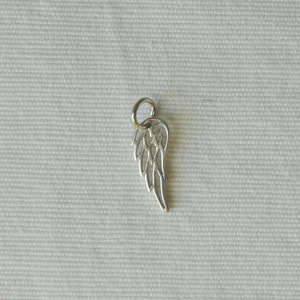May include: Silver charm in the shape of an angel wing. The wing is detailed and has a delicate design.
