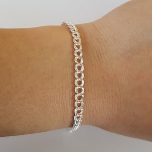 Puede incluir: Un brazalete de cadena de plata con un diseño delicado y trenzado. El brazalete se muestra en la muñeca de una persona.