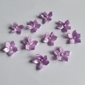 Puede incluir: Una colección de pequeñas flores de satén lavanda con un centro de perla. Las flores están dispuestas sobre una superficie blanca. Cada flor tiene cinco pétalos.