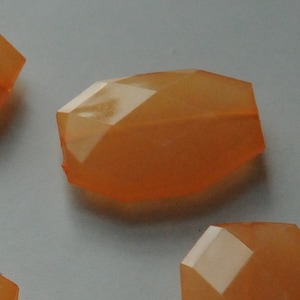 Puede incluir: Un grupo de cuatro cuentas de vidrio facetadas de color naranja. Las cuentas son translúcidas y tienen un ligero brillo iridiscente.