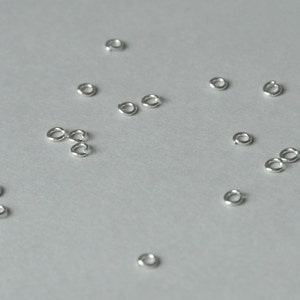 Könnte beinhalten: Eine Sammlung silberner Sprungringe, verstreut auf einer weißen Oberfläche. Die Ringe sind klein und rund, mit einer glatten, polierten Oberfläche.