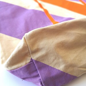 May include: Close-up of a canvas tote bag with a geometric design. The bag features a beige handle and a base with a purple stripe. The main body of the bag has a pattern of purple, white, and orange stripes.