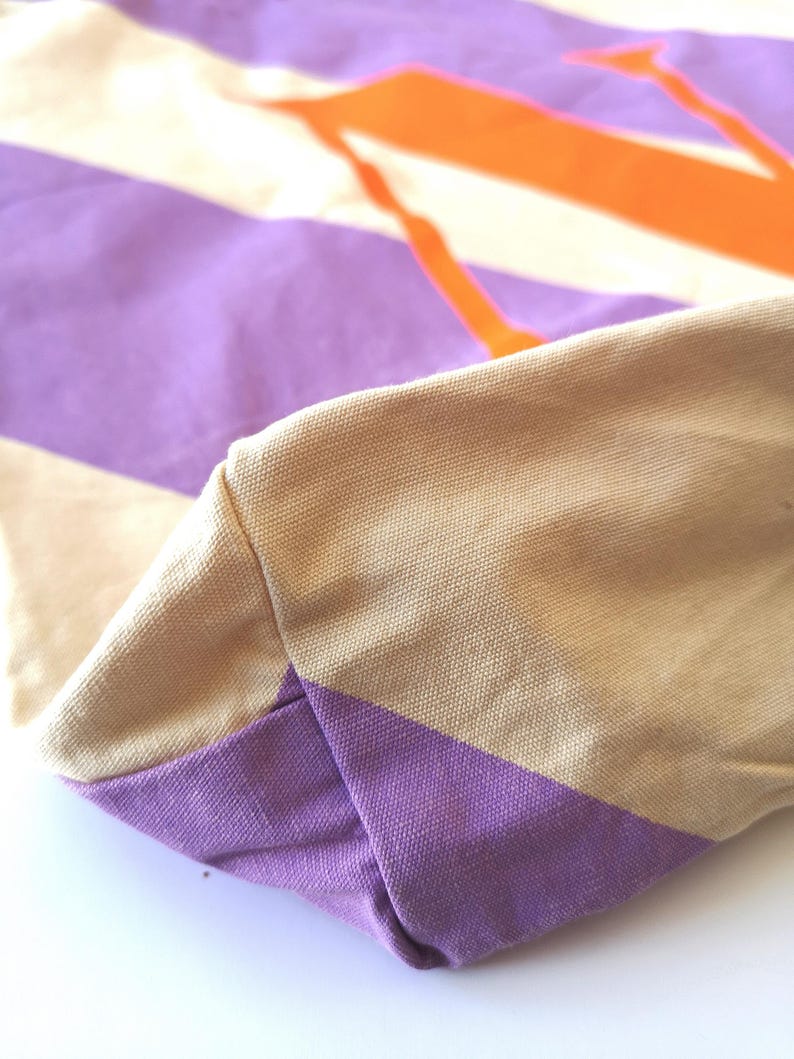 May include: Close-up of a canvas tote bag with a geometric design. The bag features a beige handle and a base with a purple stripe. The main body of the bag has a pattern of purple, white, and orange stripes.