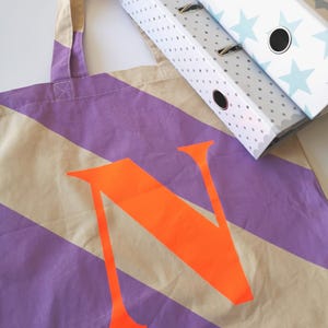 May include: A canvas tote bag with diagonal stripes in beige and purple, featuring a large orange letter "N". Two white ring binders with blue star and gray polka dot patterns are in the background.