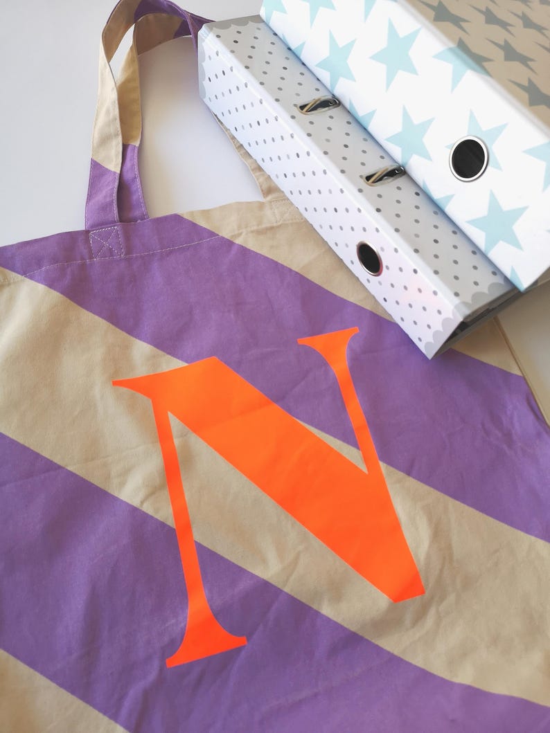 May include: A canvas tote bag with diagonal stripes in beige and purple, featuring a large orange letter "N". Two white ring binders with blue star and gray polka dot patterns are in the background.