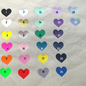 May include: A collection of heart-shaped stickers in various colors, each with a number from 1 to 26. Colors include black, white, gold, pink, orange, green, blue, and purple. The stickers are arranged on a light-colored fabric.