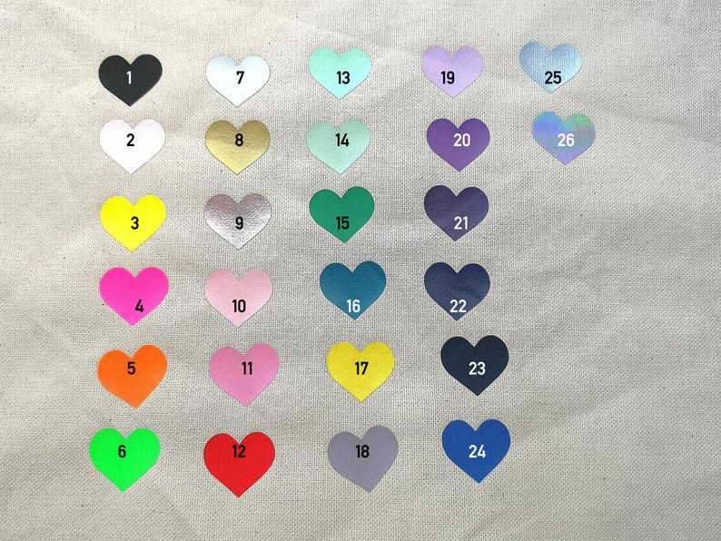May include: A collection of heart-shaped stickers in various colors, each with a number from 1 to 26. Colors include black, white, gold, pink, orange, green, blue, and purple. The stickers are arranged on a light-colored fabric.