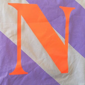 May include: Close-up of a fabric with diagonal stripes in white and lavender. A large, bright orange letter "N" is centered on the fabric. The fabric appears to be a light cotton or linen.