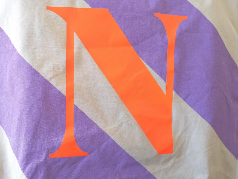 May include: Close-up of a fabric with diagonal stripes in white and lavender. A large, bright orange letter "N" is centered on the fabric. The fabric appears to be a light cotton or linen.