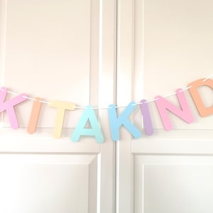 May include: A decorative banner featuring individual pastel-colored letters strung together on a white cord. The letters spell out the word KITAKIND in a sequence of pink, orange, yellow, light blue, and purple, creating a colorful hanging display.