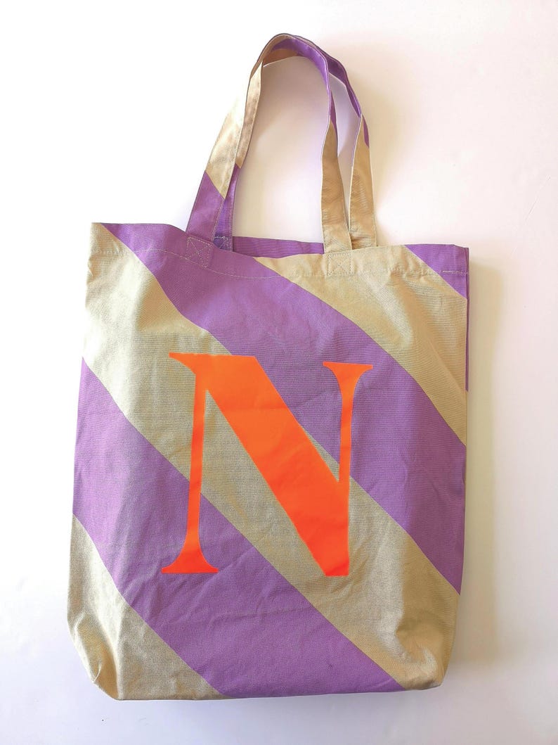 May include: A canvas tote bag with a purple and tan diagonal stripe pattern. The bag features a large orange letter "N" in the center. The tote has tan handles.