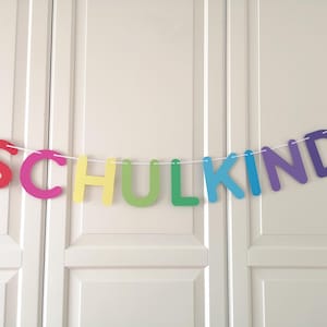 Könnte beinhalten: Ein dekoratives Banner mit dem Wort SCHULKIND, das aus einzelnen, bunten Großbuchstaben besteht. Die Buchstaben sind auf einer dünnen weißen Schnur aufgereiht und zeigen eine Abfolge in Rot, Pink, Gelb, Grün, Blau und Violett.